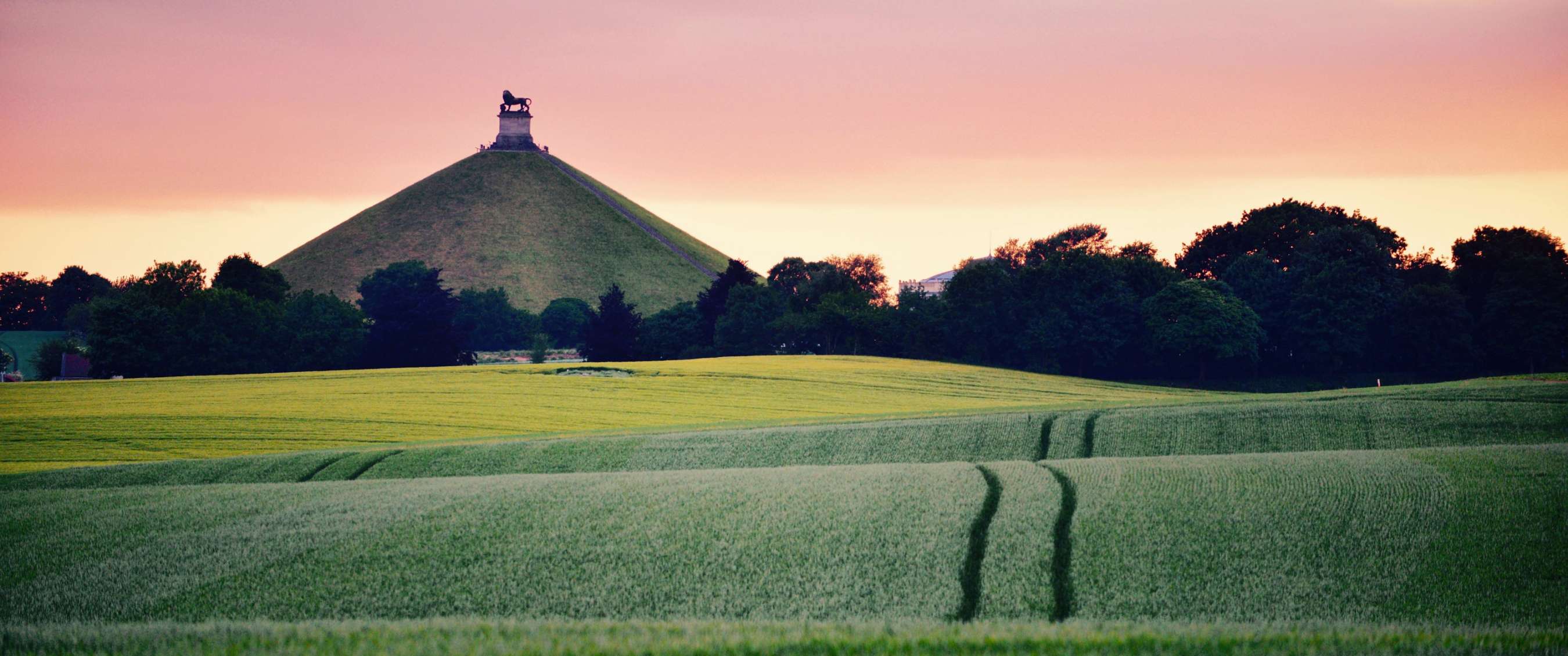 Landschap met akkers, groene heuvel en standbeeld tegen gekleurde lucht.