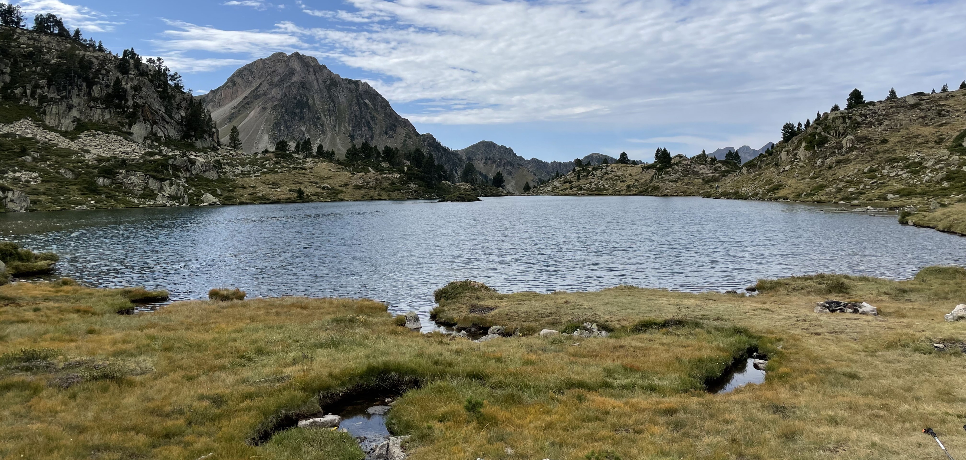 Lac de montagne entouré de pentes rocheuses, prairie au premier plan et chaîne de montagnes sous un ciel nuageux.