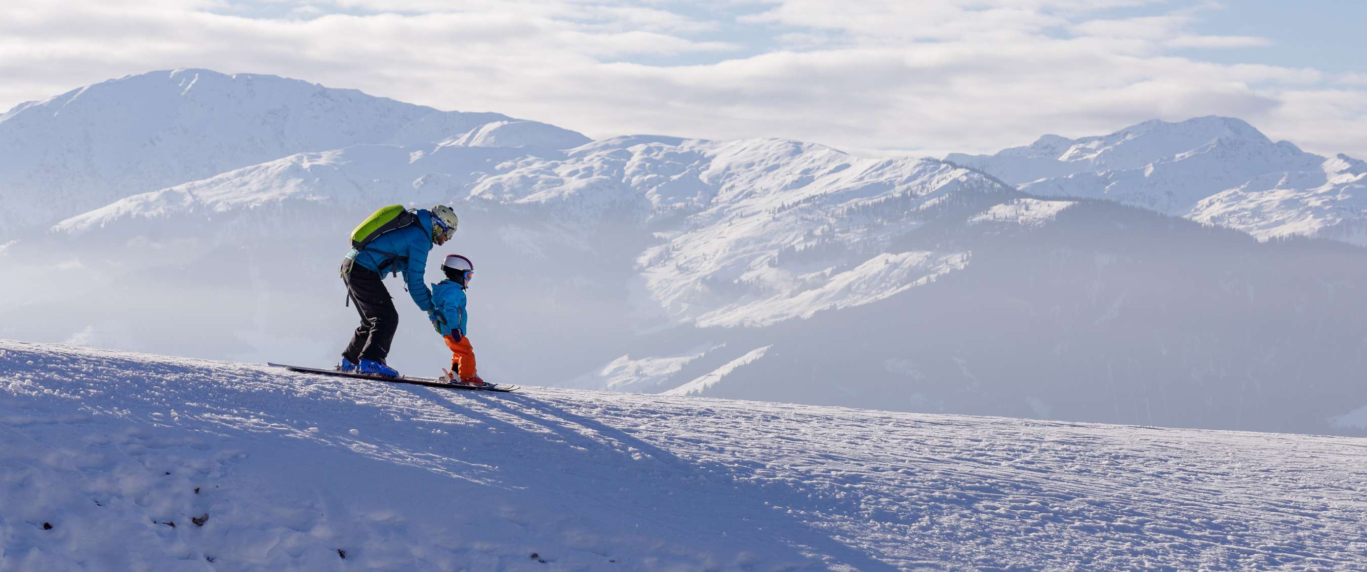 Volwassene helpt een kind skiën op een berghelling. Besneeuwde bergen op de achtergrond.