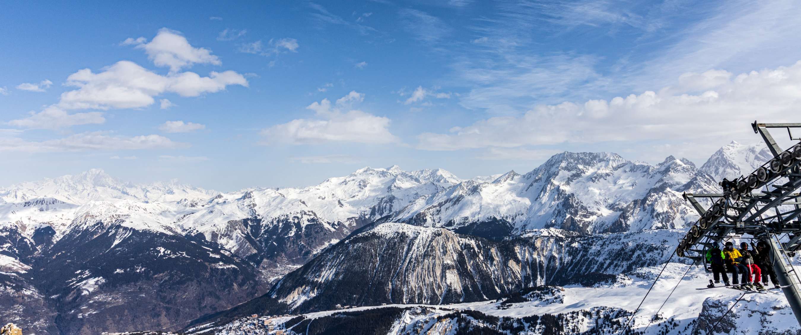 Een stoeltjeslift met passagiers op de voorgrond en daarachter besneeuwde bergen onder een helderblauwe lucht met witte wolken in Trois Vallées.